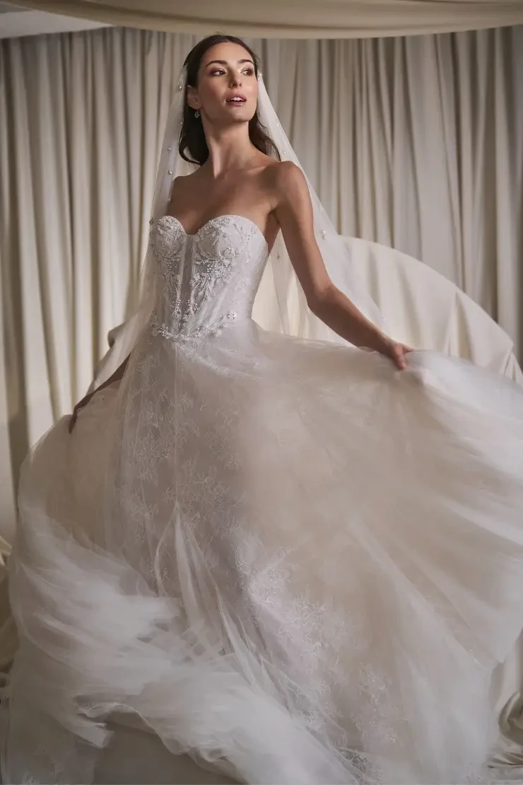 A model wearing a beautiful strapless wedding gown with intricate detailing, posing elegantly against a soft, draped background.