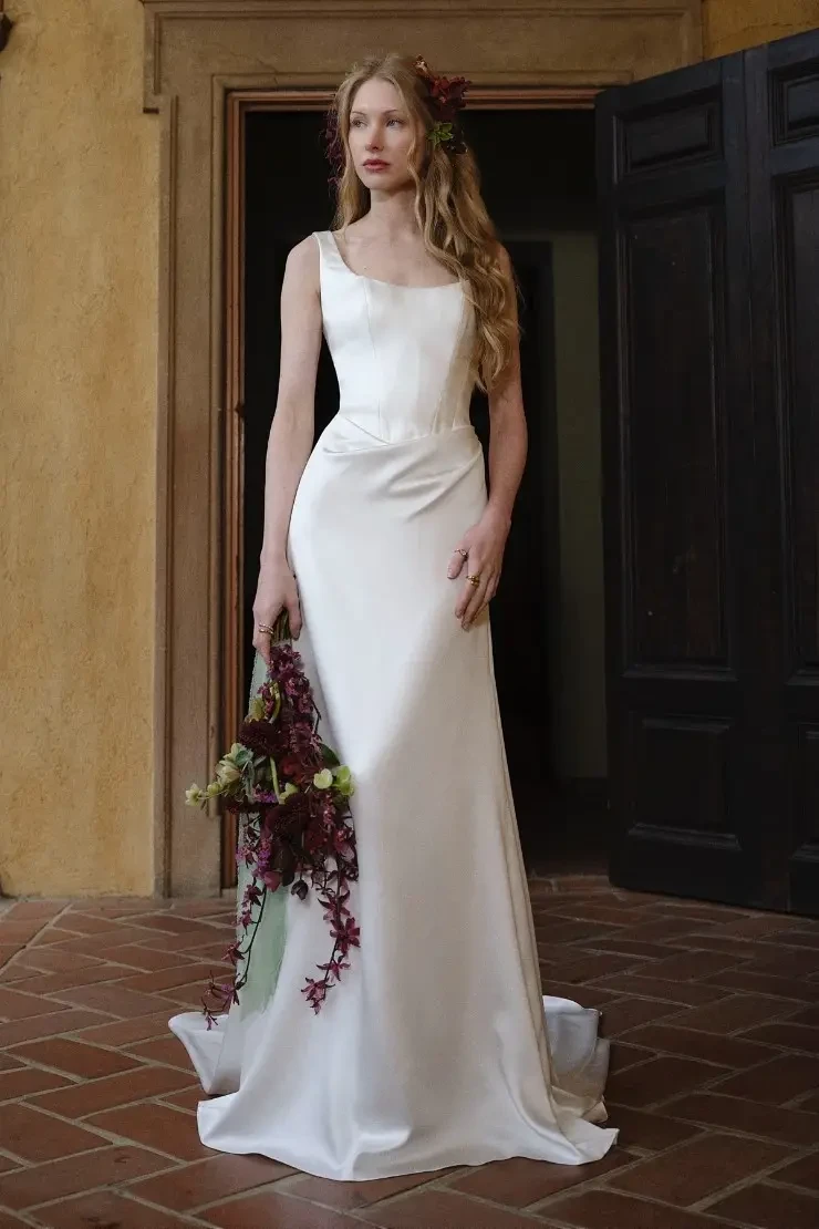 A model wearing a simple, elegant white wedding dress stands in a doorway, holding a bouquet of flowers.