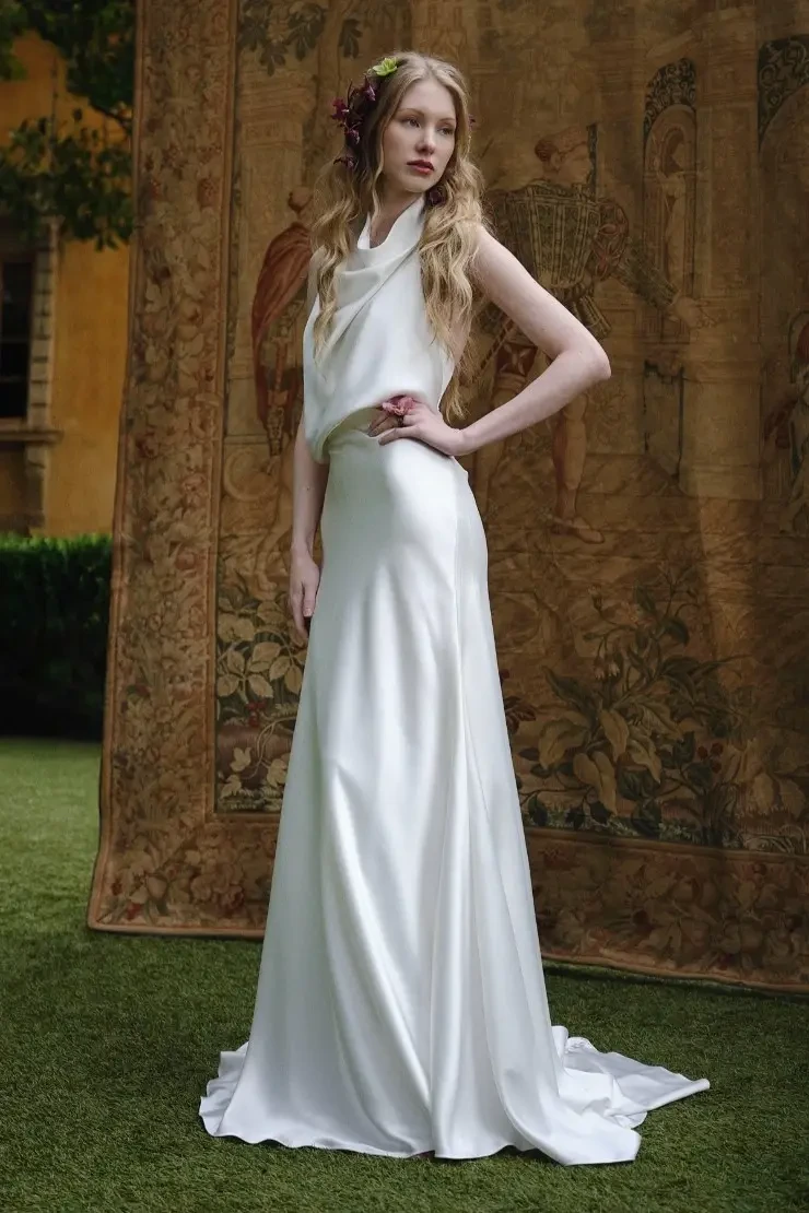 A model wearing a sleek, white gown stands in front of a vintage tapestry backdrop. The gown features a draped top and a fitted skirt, with the model's long hair styled elegantly.